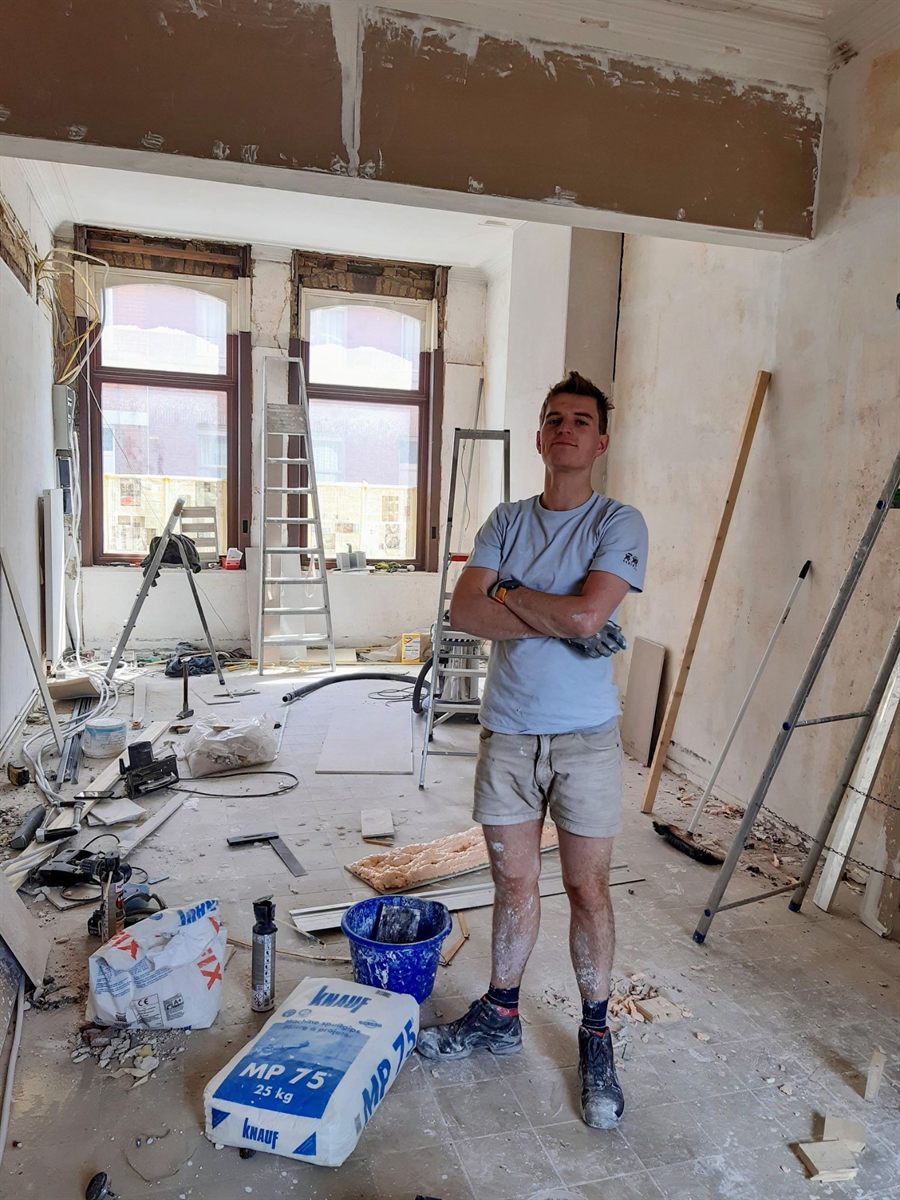 Emiel met gekruiste armen in een kamer vol ladders en bouwmateriaal tijdens de verbouwing